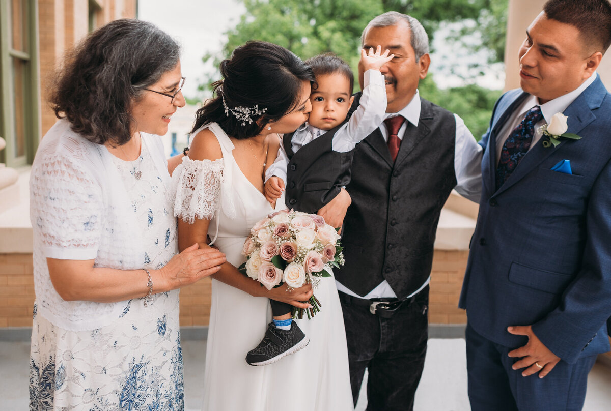 Couples Photography,, woman in a wedding dress is holding a toddler boy as an older woman, older man, and young man smile and look at him.
