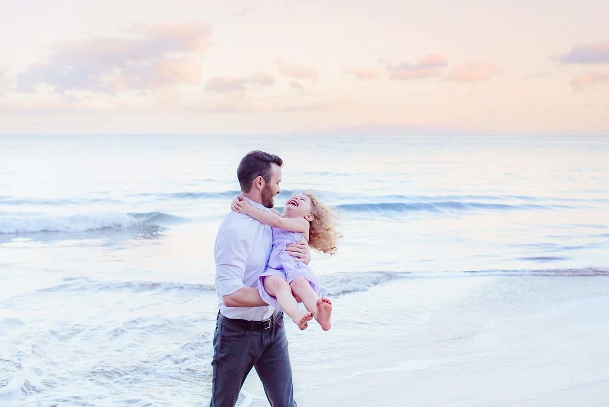 Happy dad holds and spins his daughter during a sunrise Maui portrait shoot next to the ocean in Wailea wit Love + Water