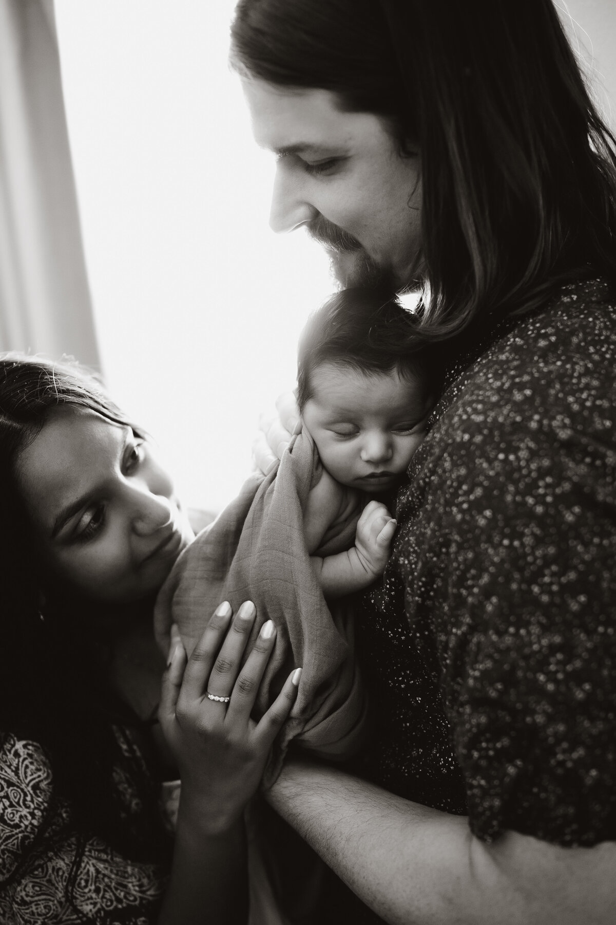 a black and white portrait of a family of 3 with a  newborn baby standing and hugging