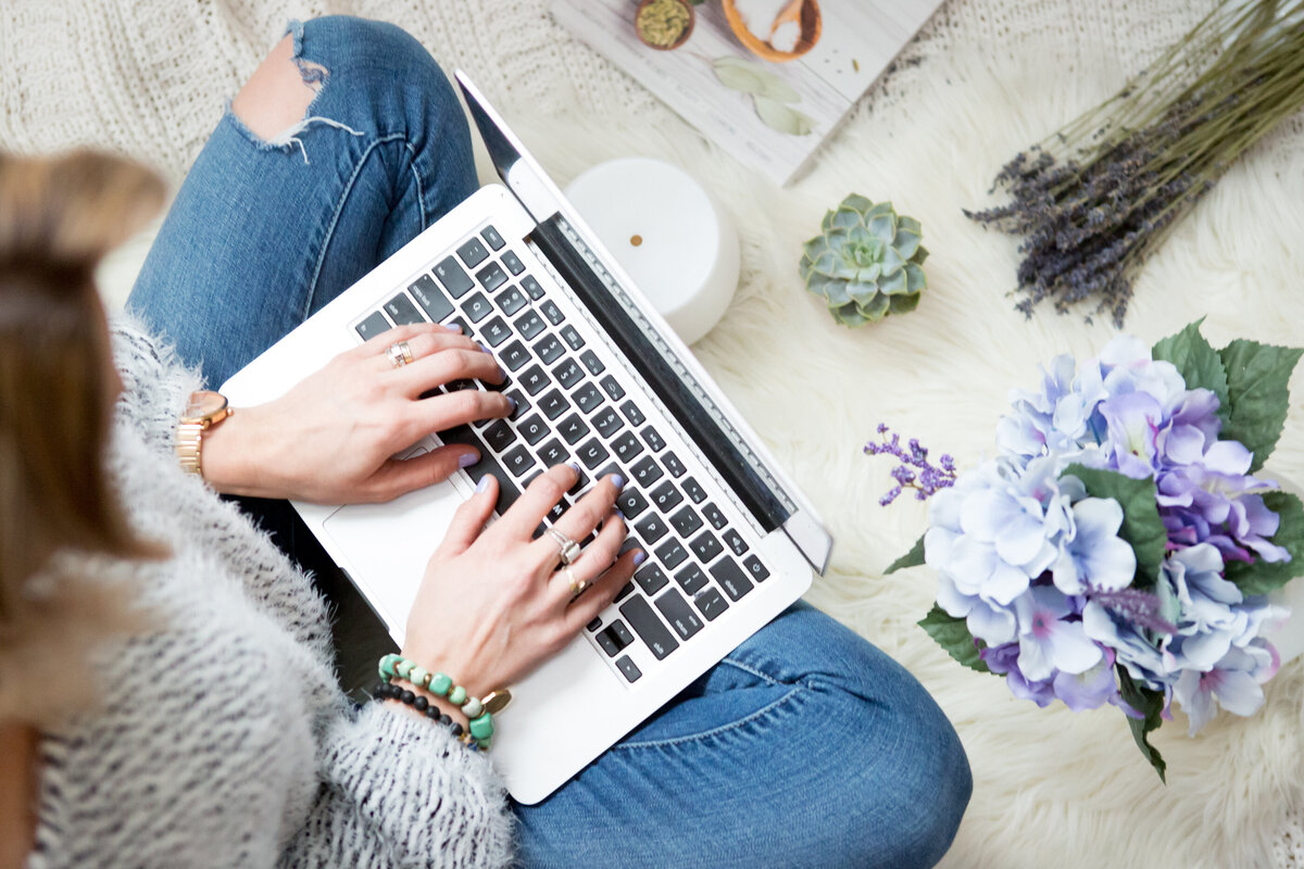 detail shot of white woman in ripped blue jeans typing on a laptop with a blue bouquet, lavender and succulent sitting on a white furry blanket New York branding photographer Milestone Images