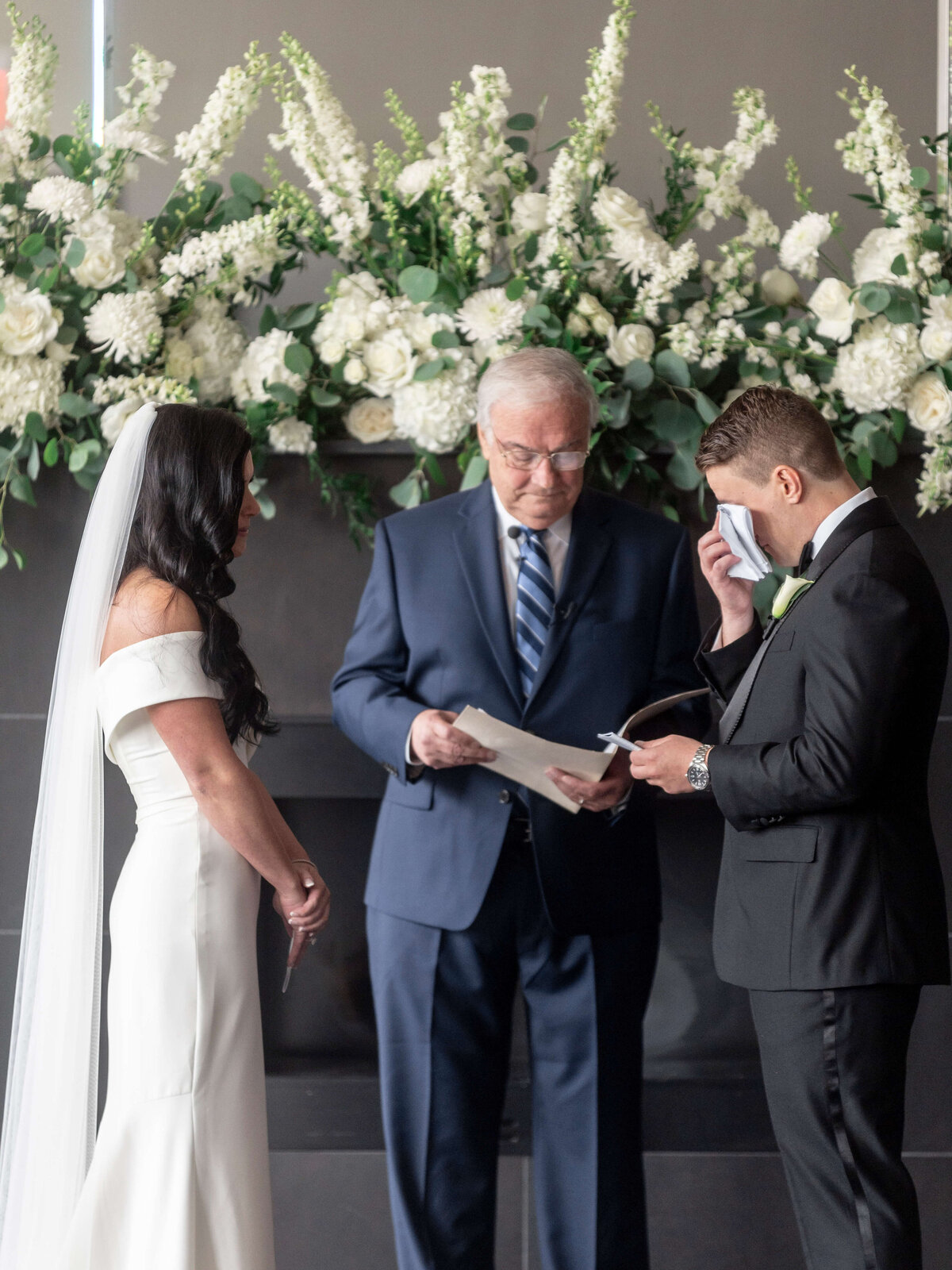A couple stands at their wedding ceremony with an officiant. The bride, in a white dress and veil, faces the groom in a black suit. The groom wipes a tear with a handkerchief. Lush white flowers and greenery decorate the background.