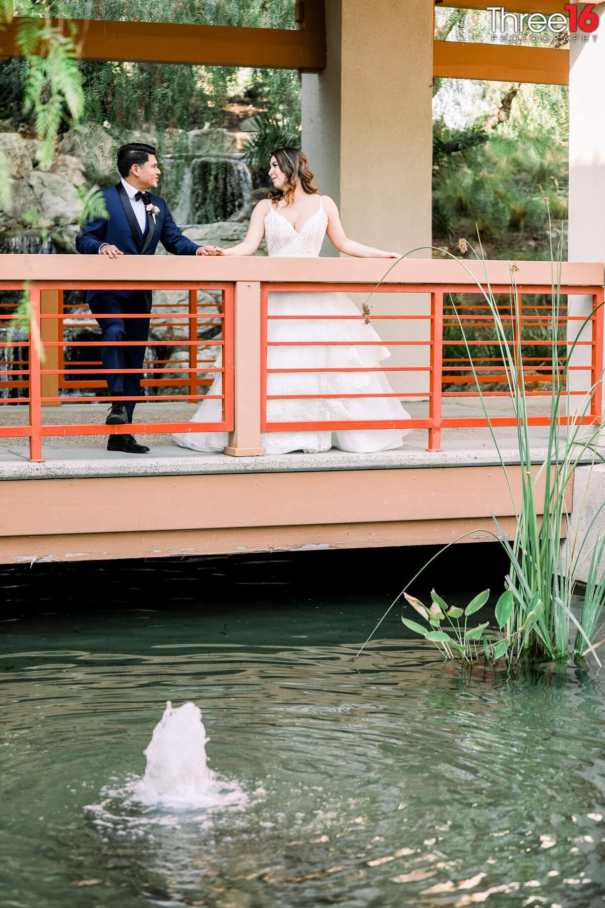 Bride and Groom stand against a rail overlooking a pond with water shooting out of the pond