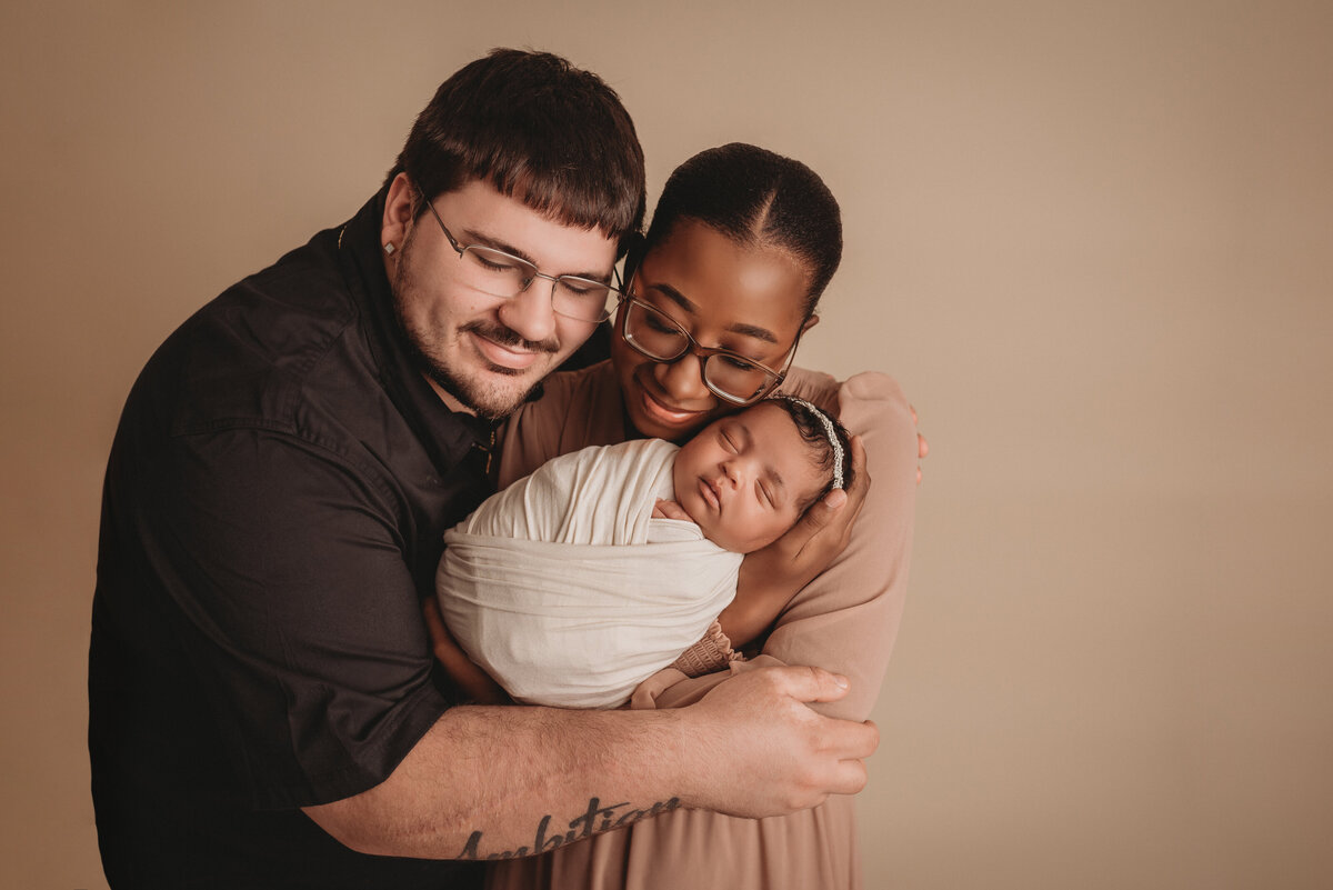 Newborn family portrait at Marietta, GA newborn photography studio with mom, dad and baby girl wearing blush pink, cream and black on a tan backdrop