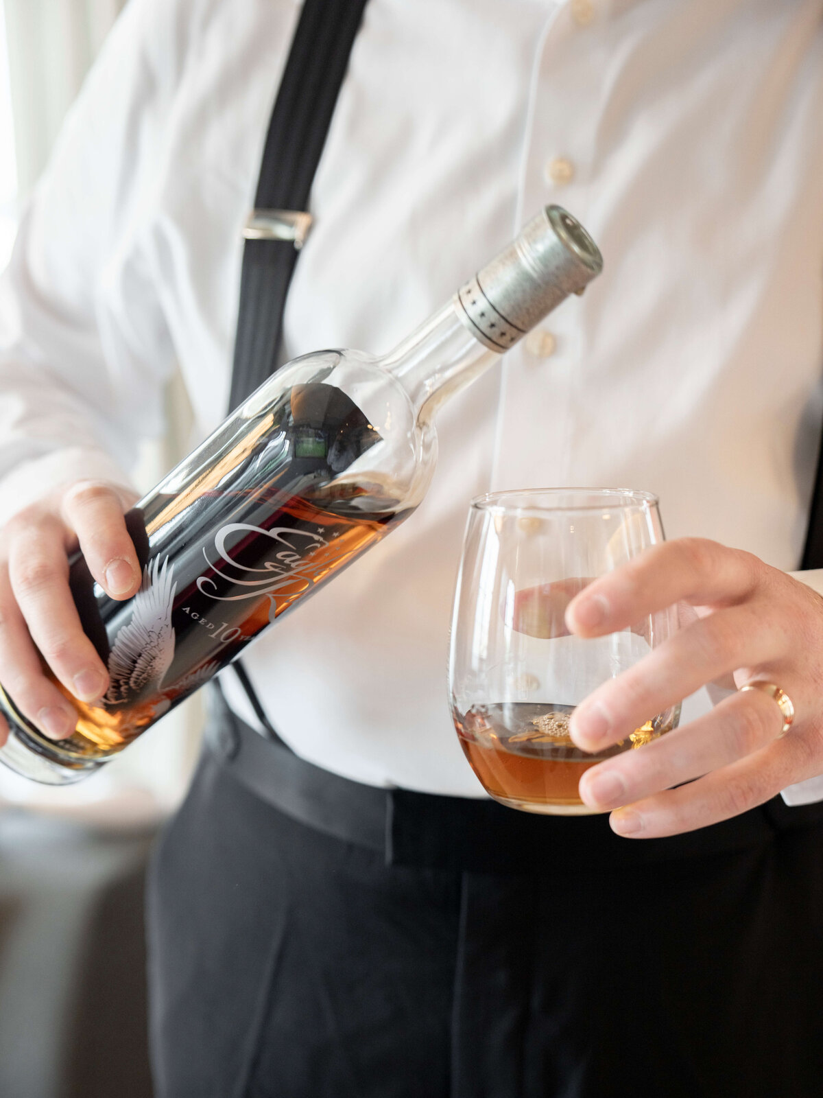 A person in a white shirt and black suspenders is pouring whiskey from a bottle into a glass. The bottle features a label with a feather design. The person has a wedding ring on their left hand.