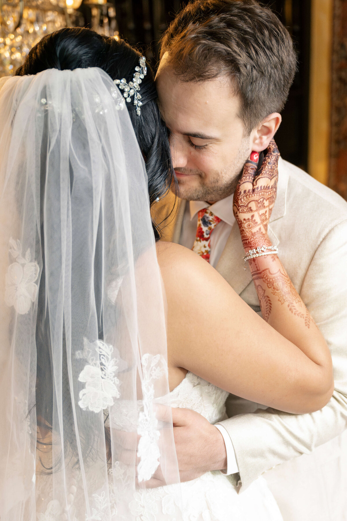 A bride and groom embrace intimately on their wedding day. The bride wears a white gown with a lace veil and has intricate henna art on her hands. The groom is in a light suit with a floral tie, both sharing a tender moment.