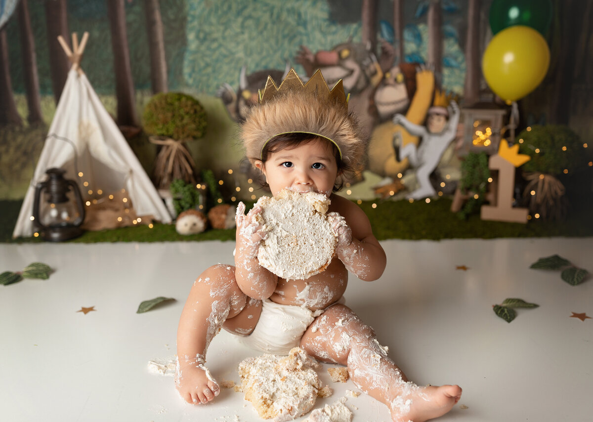 "Where the Wild Things Are" themed cake smash at top West Palm Beach cake smash photographer. Baby boy is sitting in white bottoms covered in cake. He is holding a cake round in both hands, bringing it up to his face to eat. He is wearing a fur-trimmed crown and looking at the camera. In the background, there is the a "Where the Wild Things Are" backdrop, teepee, and forest decor.