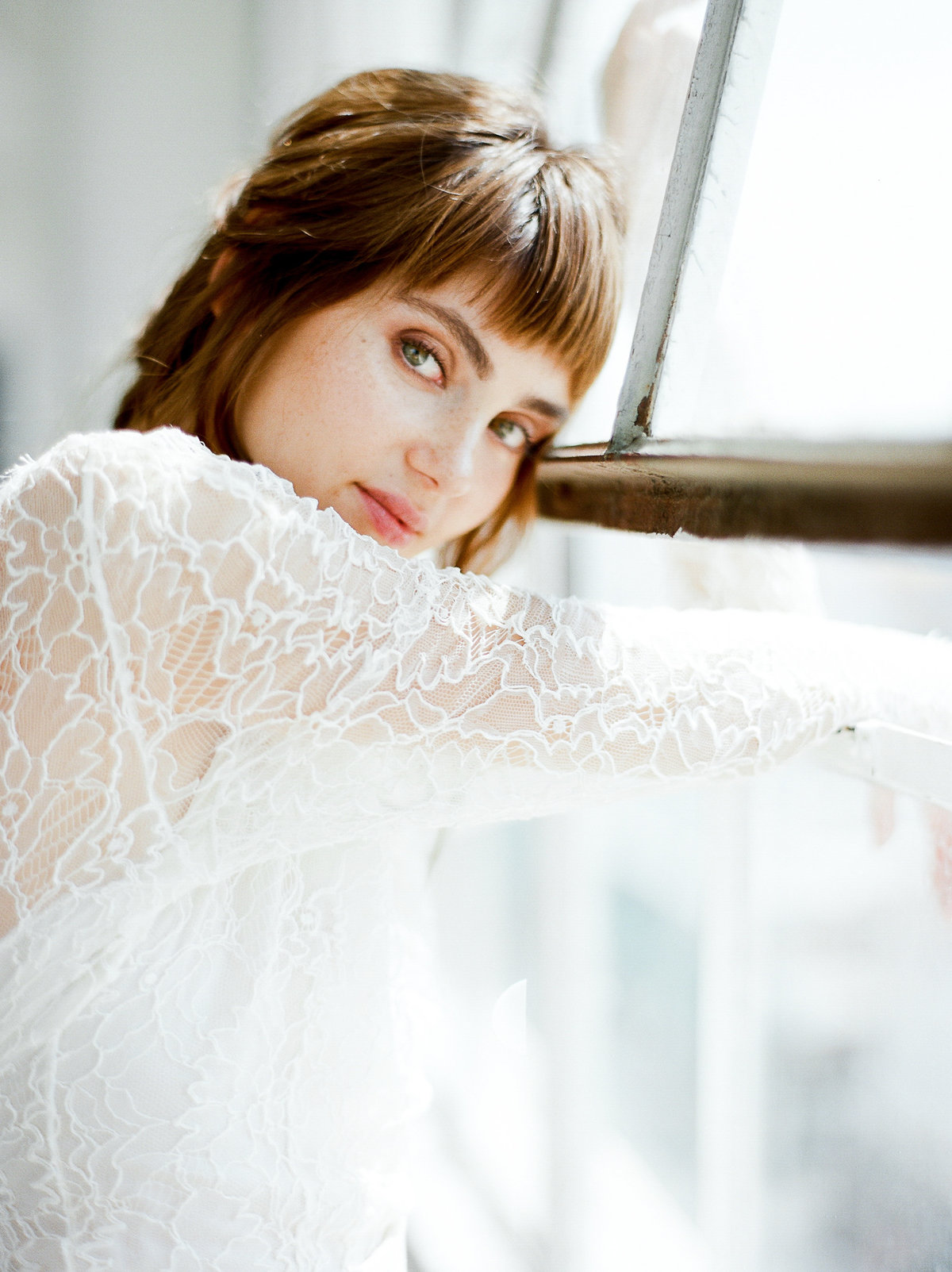 bride leaning on a window looking at camera with green eyes and brown hair and a lace cortana gown on