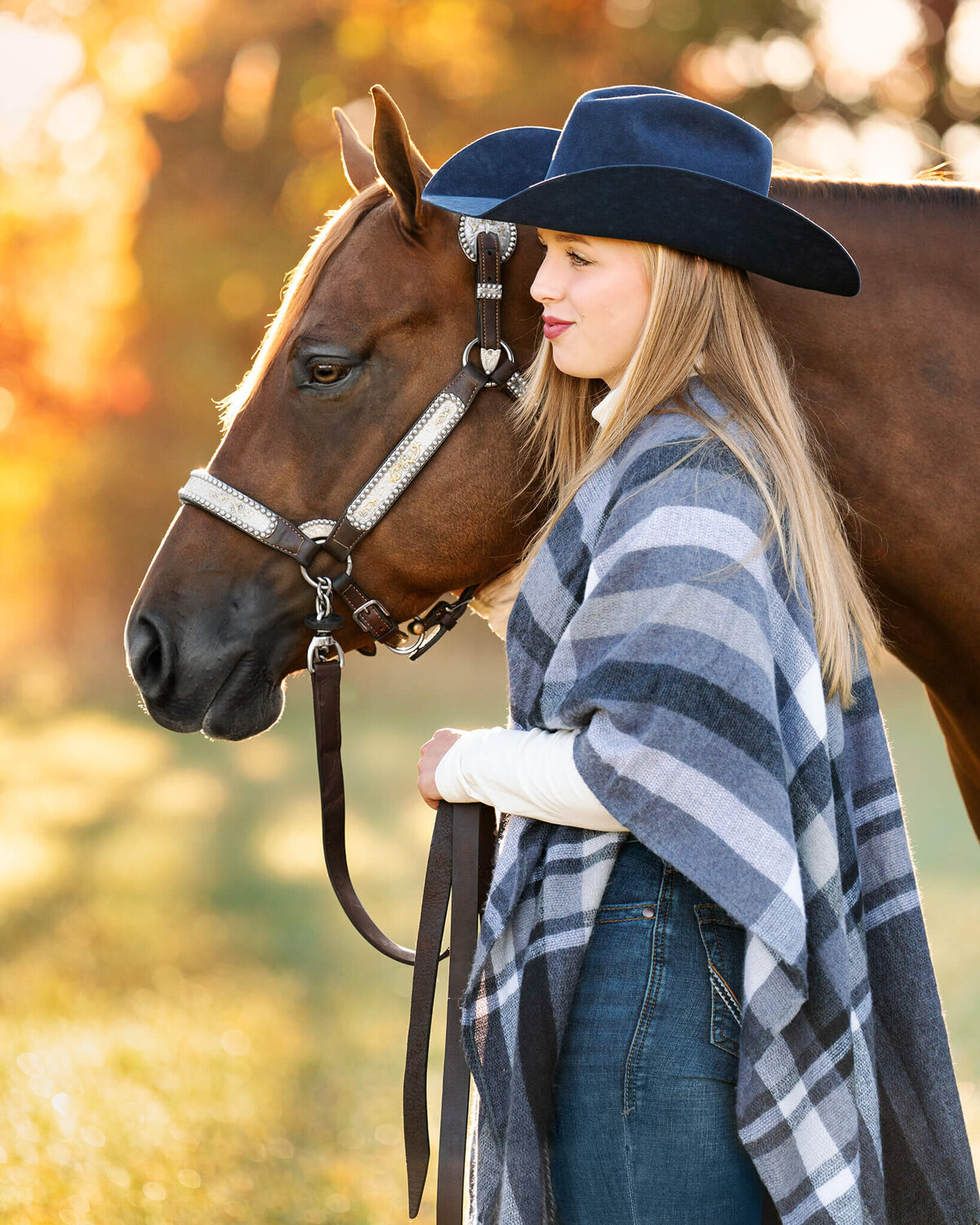 blonde equestrian wearing a navy blue cowboy hat and a blue and white stripped shaw standing next to her chestnut paint horse. they are both looking off camera so we see their profile. in the back is yellow and orange fall leaves.