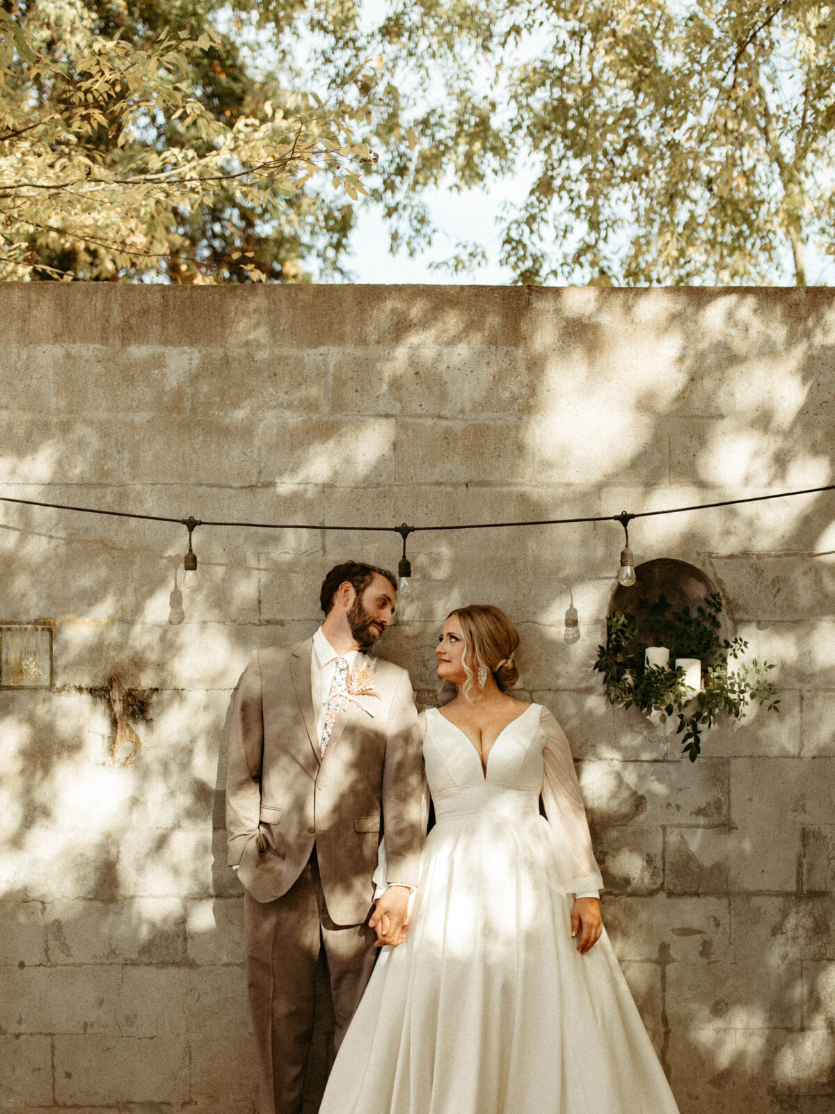 Bride and groom in wedding attire standing side by side leaning against wall with string lights hanging over them