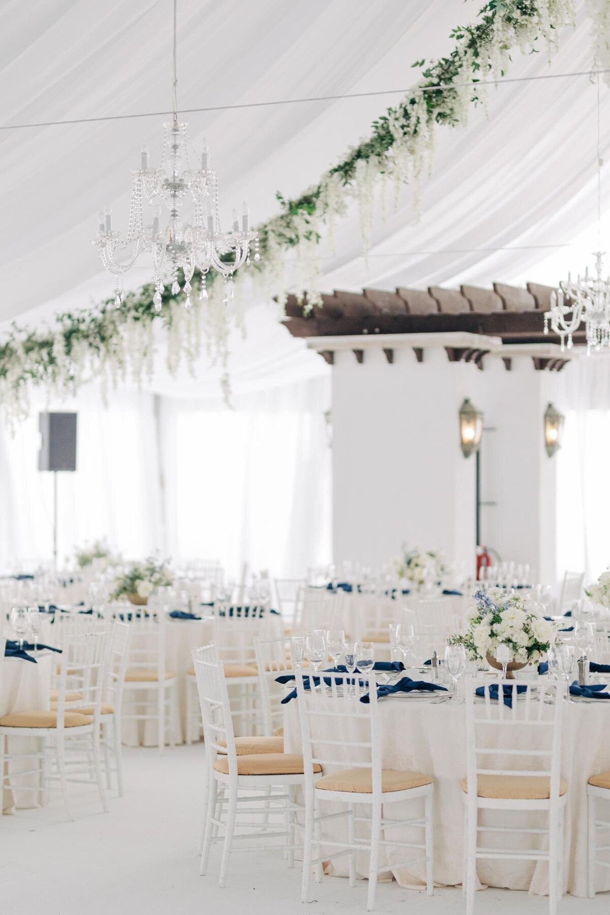 Elegant wedding reception setup inside a white tent with chandeliers, white chairs with blue ribbons, and floral decorations.