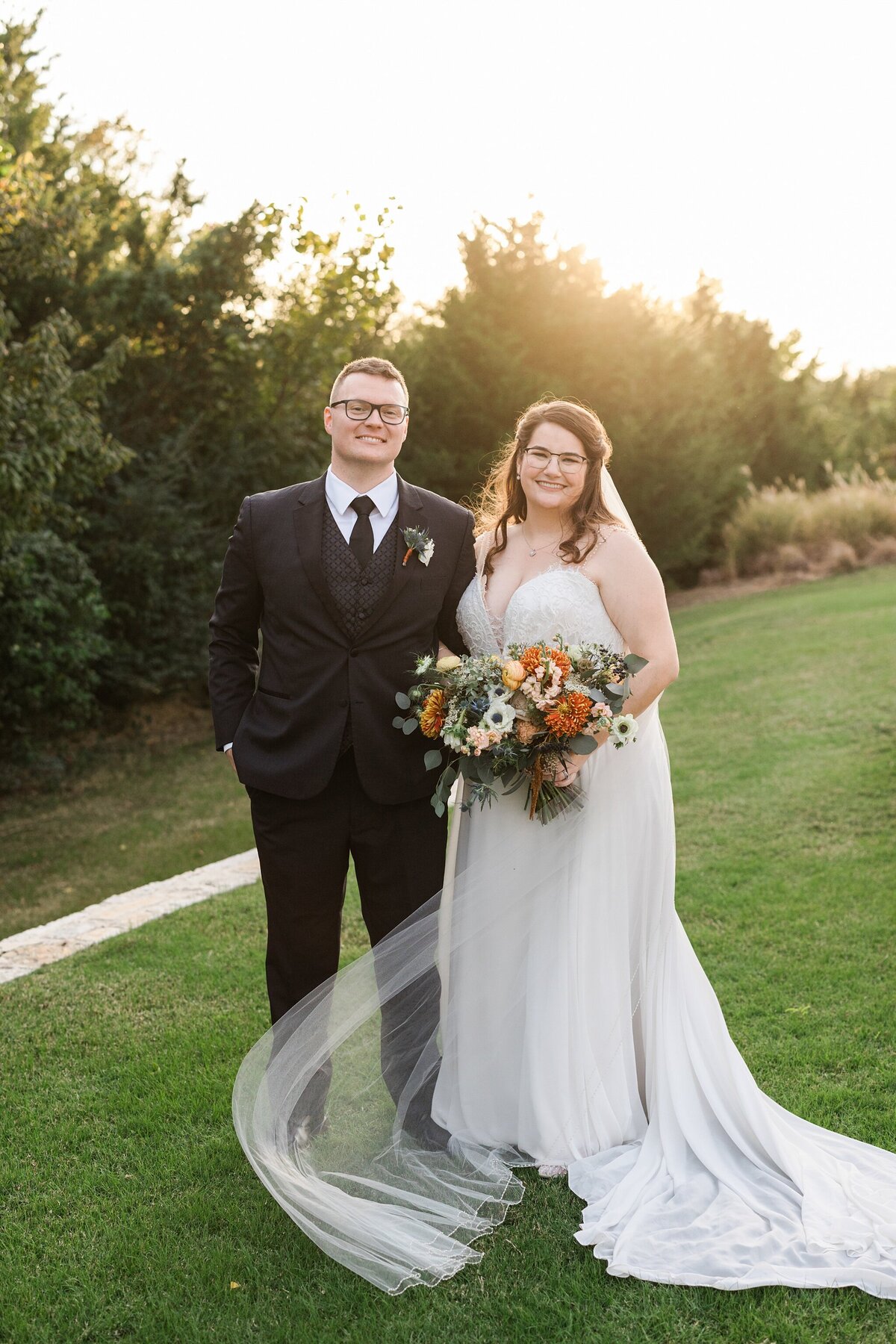 A portrait of a bride and groom posing together outside after their wedding ceremony at The Laurel in Grapevine, Texas. The bride is on the right and is wearing a long, flowing, sleeveless, white dress with a long, flowing veil and is holding a large, colorful bouquet. The groom is on the left and is wearing a black, three piece suit with a boutonniere.