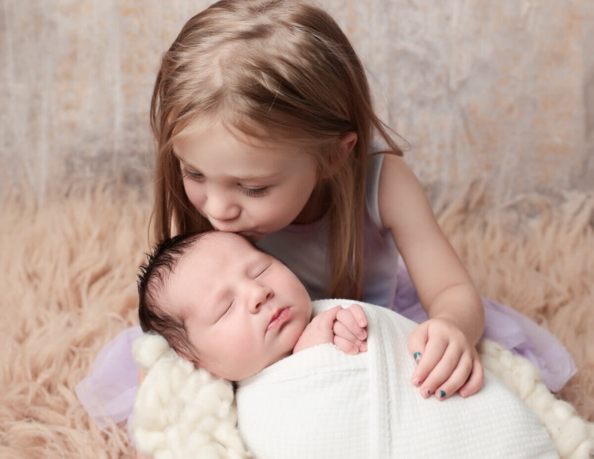 A heartwarming photograph of a sibling leaning down to kiss their newborn brother in a cozy photography studio in Rochester, NY. The soft lighting and warm colors capture the love and connection between the siblings, highlighting the joy of welcoming a new family member.