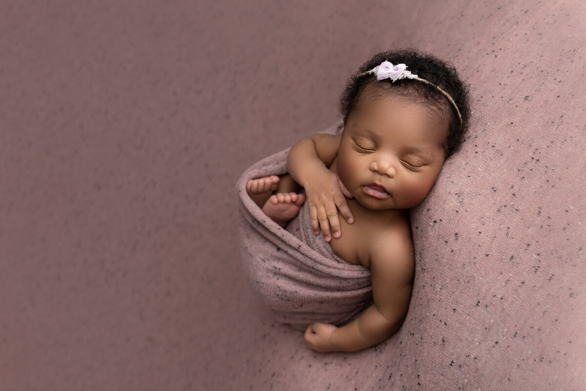 Philadelphia's best newborn photographer, Katie Marshall captures a fine art newborn image. Black baby girl is swaddled in mauve with her feet folded up and toes peeking out. One hand is draped atop of her chest. She is wearing a delicate headband and has lots of hair.