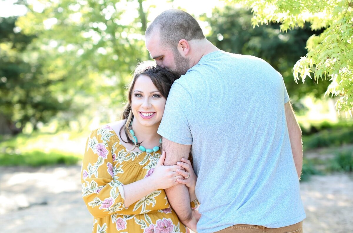 A man kissing his fiancee's head while she's smiling for the camera during an engagement session in Raleigh, NC. Raleigh engagement photographer.