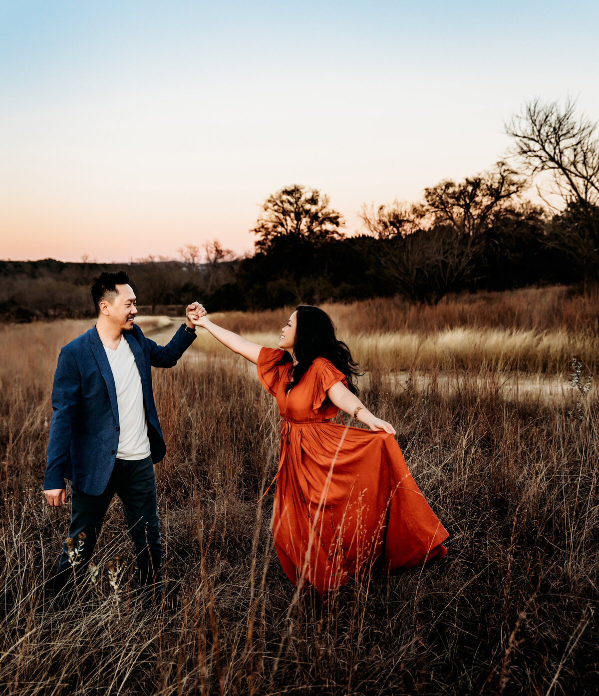 Couples Photography, Man in a blue blazer holding hands with a woman wearing a rust dress, twirling her in a field.