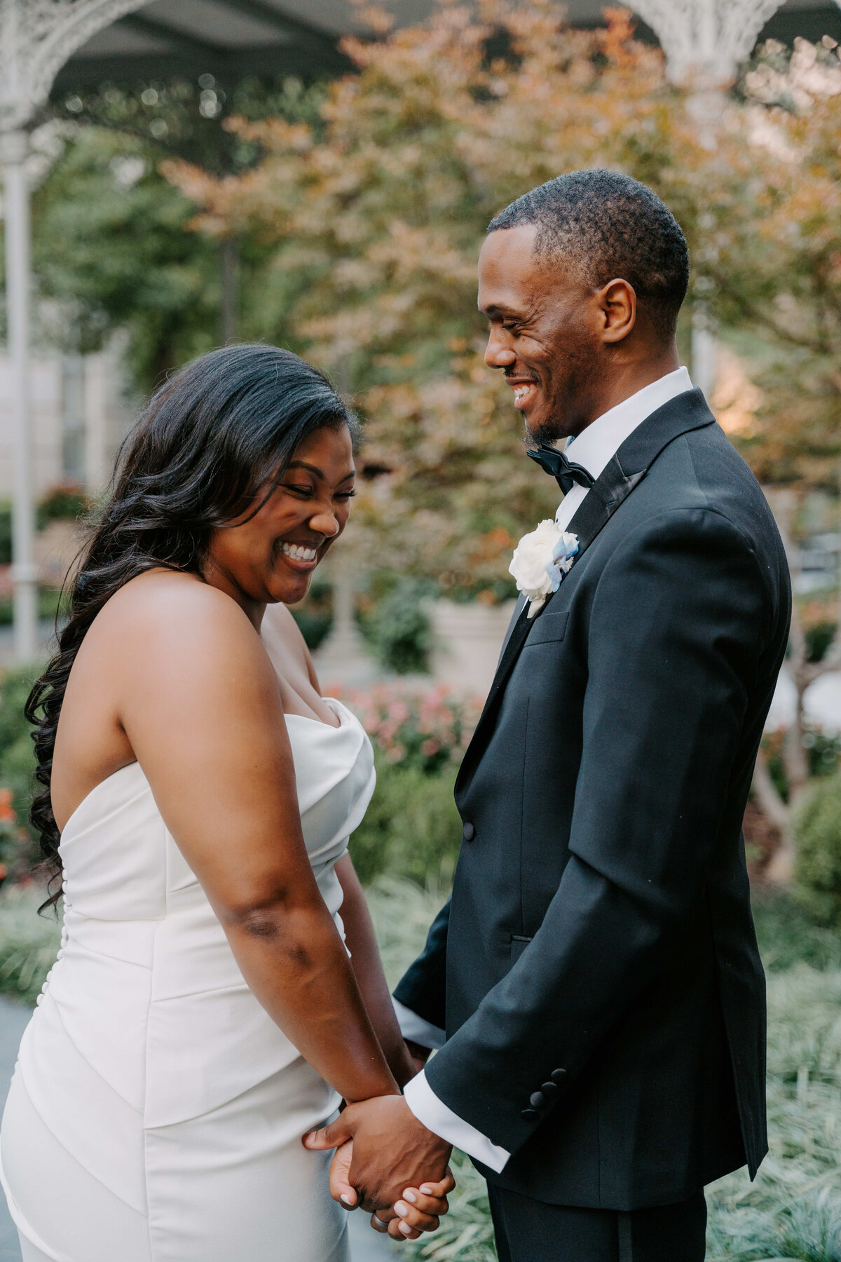 A joyful couple in wedding attire holding hands outdoors. The bride in a white dress and the groom in a black suit and bow tie, both smiling warmly at each other.