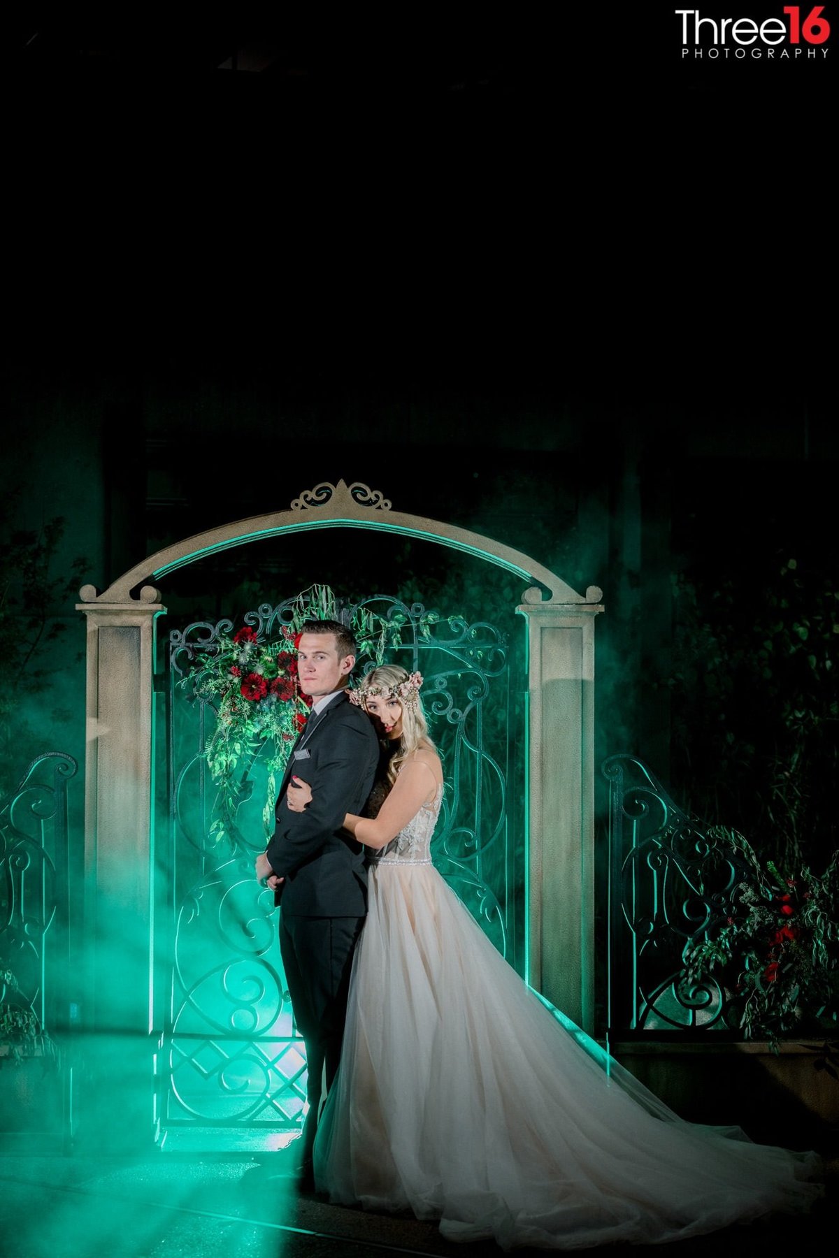 Bride embraces her Groom from behind as they pose at night in front of a Center Club Orange County gate with a greenish color in the background