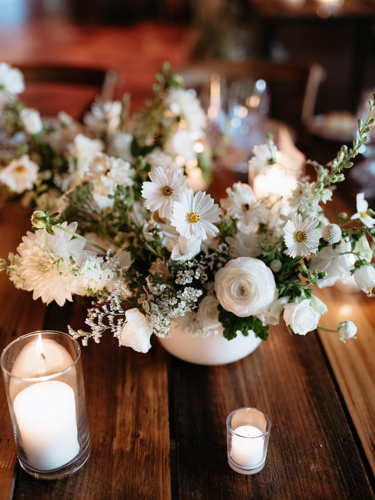 Closeup of romantic white daisy and ranunculus bouquet surrounded by white candles on deep brown farmhouse table