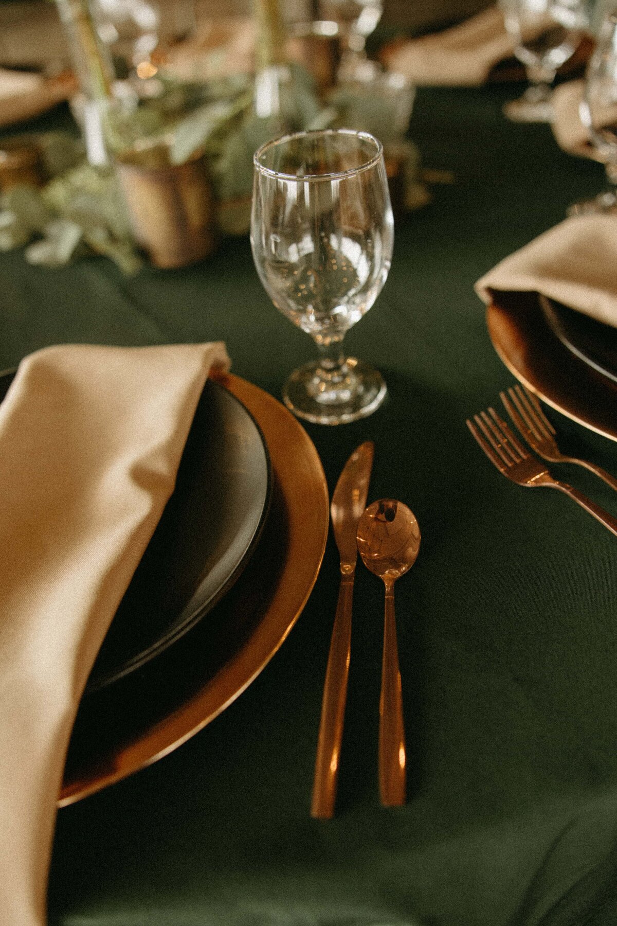 Elegant wedding table setting featuring a dark green tablecloth, a rustic brown plate with a gold-rimmed napkin, and coordinating cutlery next to a clear water glass.
