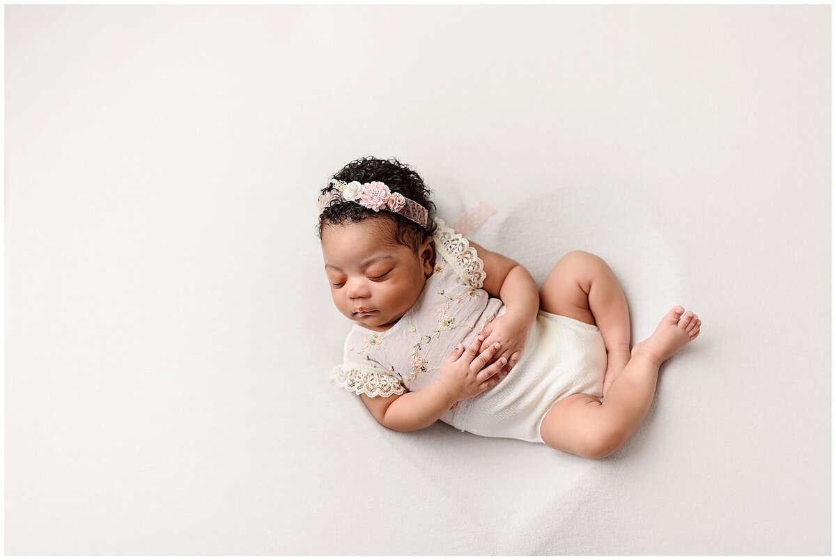 A newborn baby girl wearing a delicate one piece outfit cuddled up resting on a soft white surface