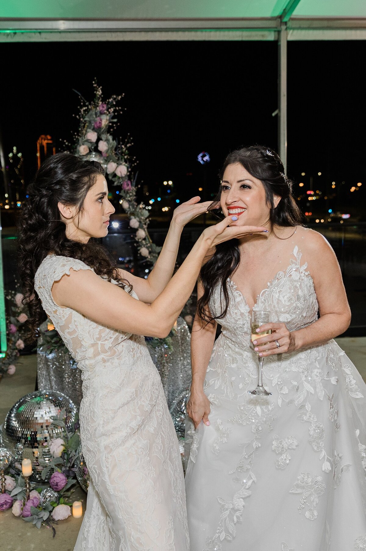 A photo of one bride playfully framing another bride's face during their wedding reception in Dallas, Texas. The bride on the left is wearing a short sleeve, intricate, white dress. The bride on the right is wearing a sleeveless, long, flowing, intricate, white dress and is holding a glass of champagne.