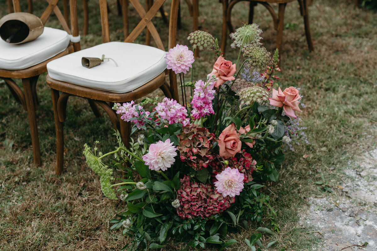Romantic ceremony aisle floral arrangement with mauve roses, pink dahlias, hydrangea, and garden greenery lining the outdoor wedding seating.