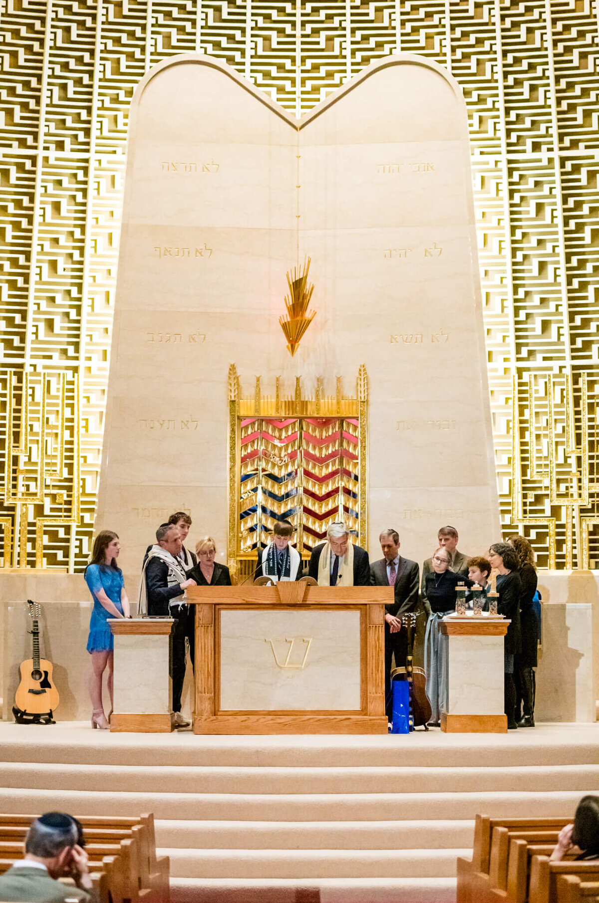A teenage boy stands at the bimah surrounded by family and rabbi reading from the torah