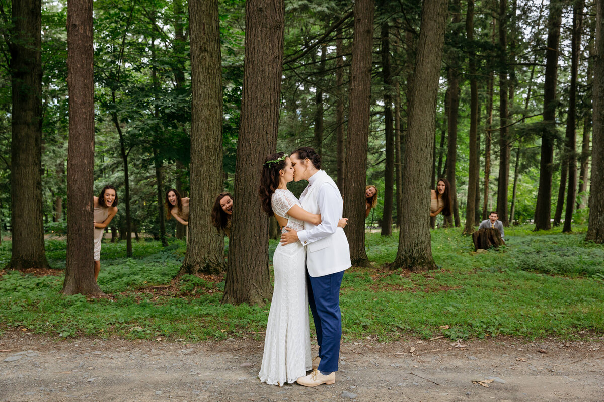 A wedding couple kissing while their wedding parties peek their heads out from trees behind the couple.