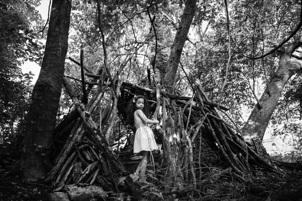 A young girl standing in a small fort made of tree branches in the woods.