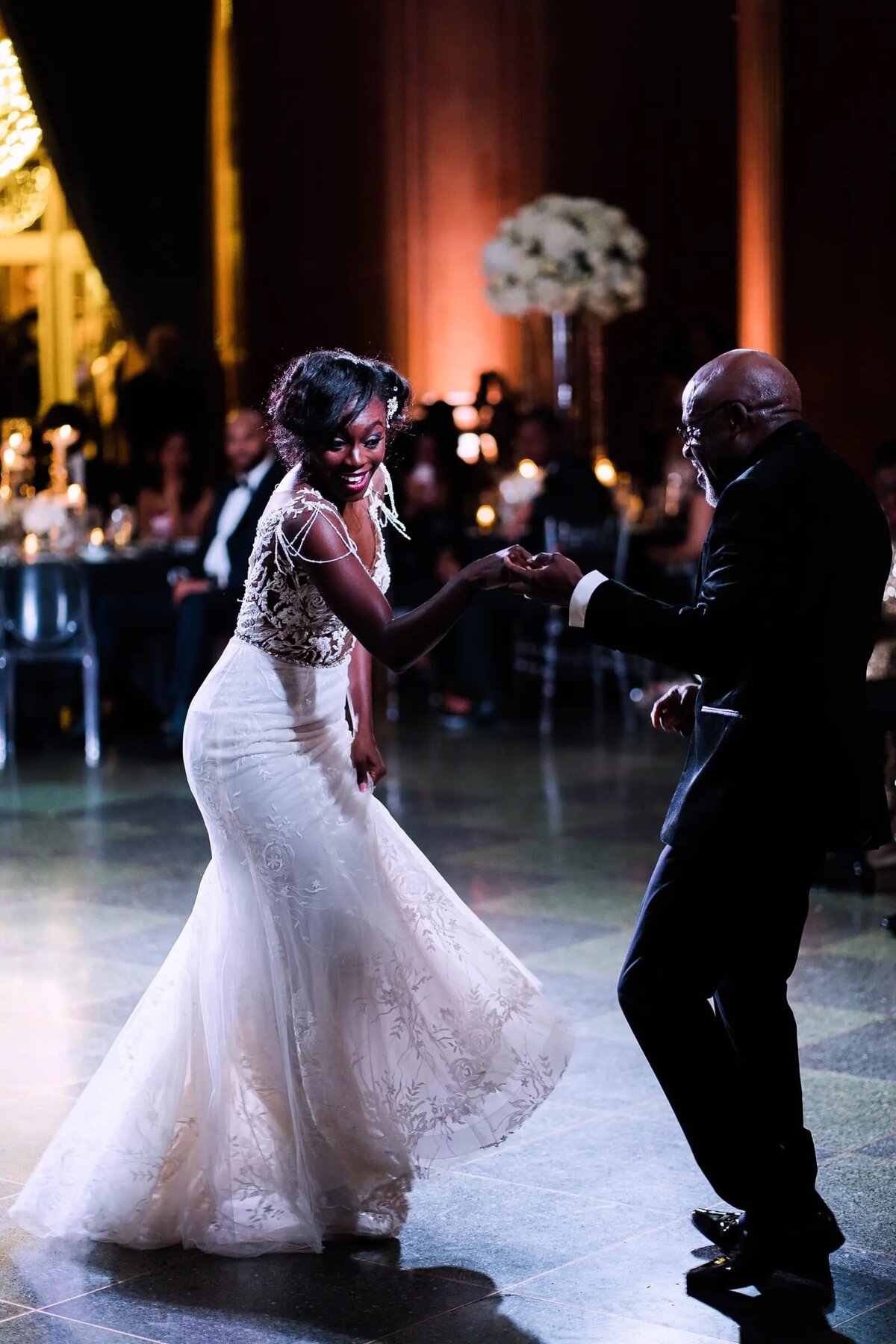 Bride dancing with an older man in a suit, guests seated in the background, at an elegantly lit wedding reception.