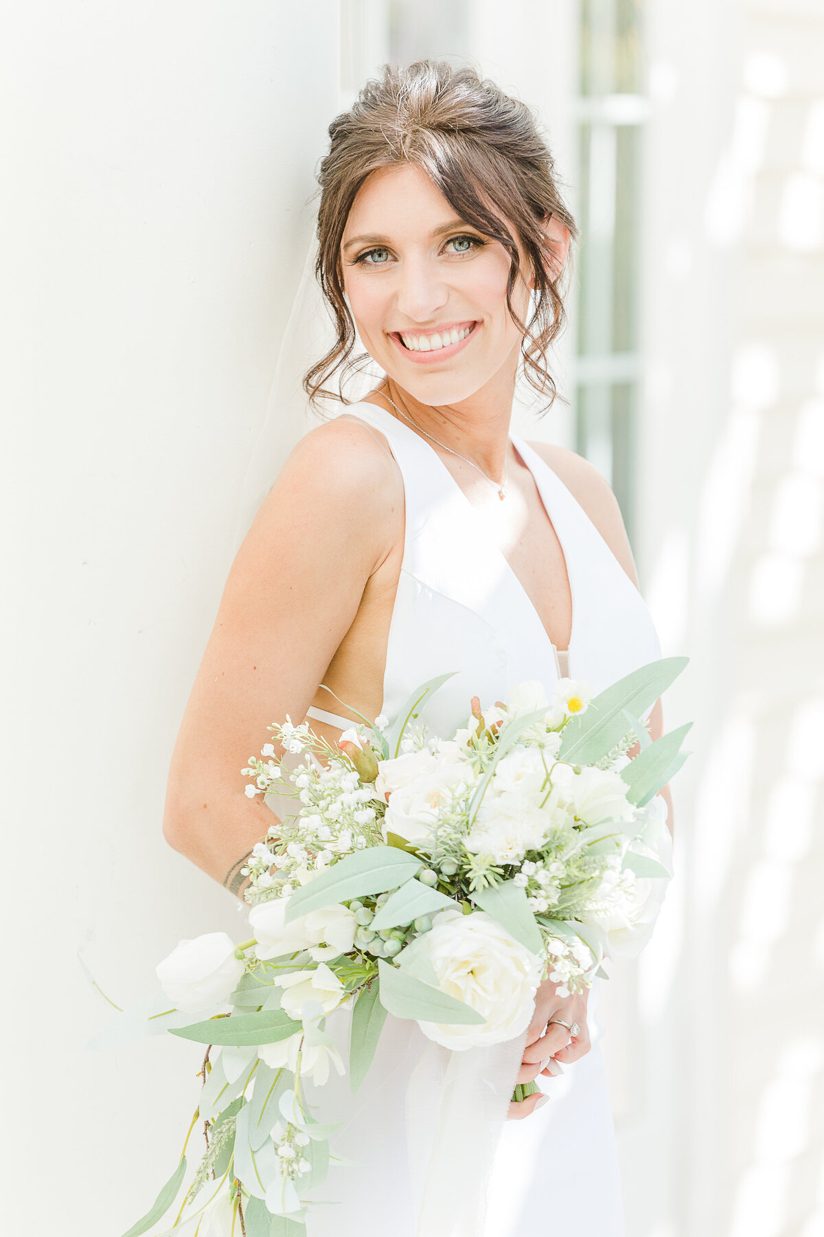 A stunning formal bridal portrait. The bride is standing angled to the camera, looking over her shoulder and smiling. She is casually holding her bouquet in front of her. Captured by best Rhode Island editorial wedding photographer Lia Rose Weddings.