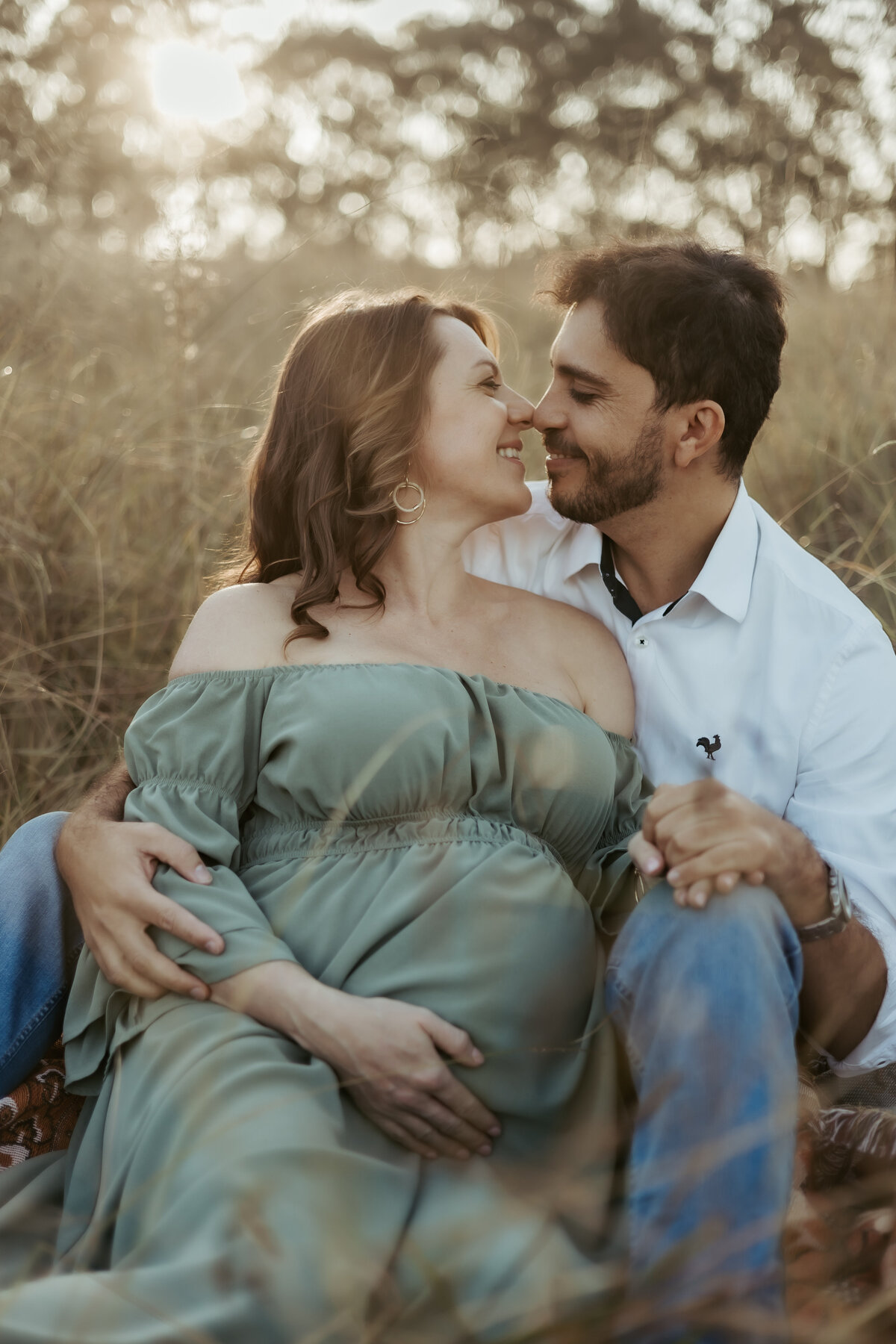 Excited parents-to-be sitting together in a gorgeous outdoor location for their maternity photoshoot smiling and their noses touching looking so happy