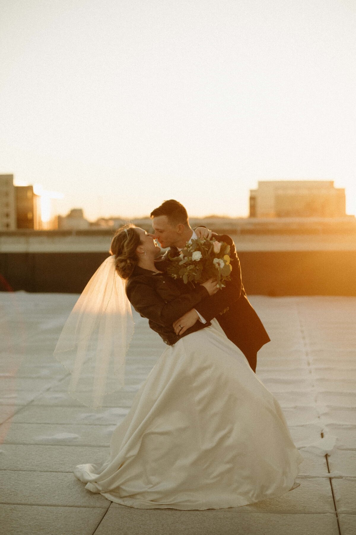 A bride and groom embrace and kiss on a rooftop at sunset in Davenport, with soft sunlight illuminating them and the city skyline in the background.