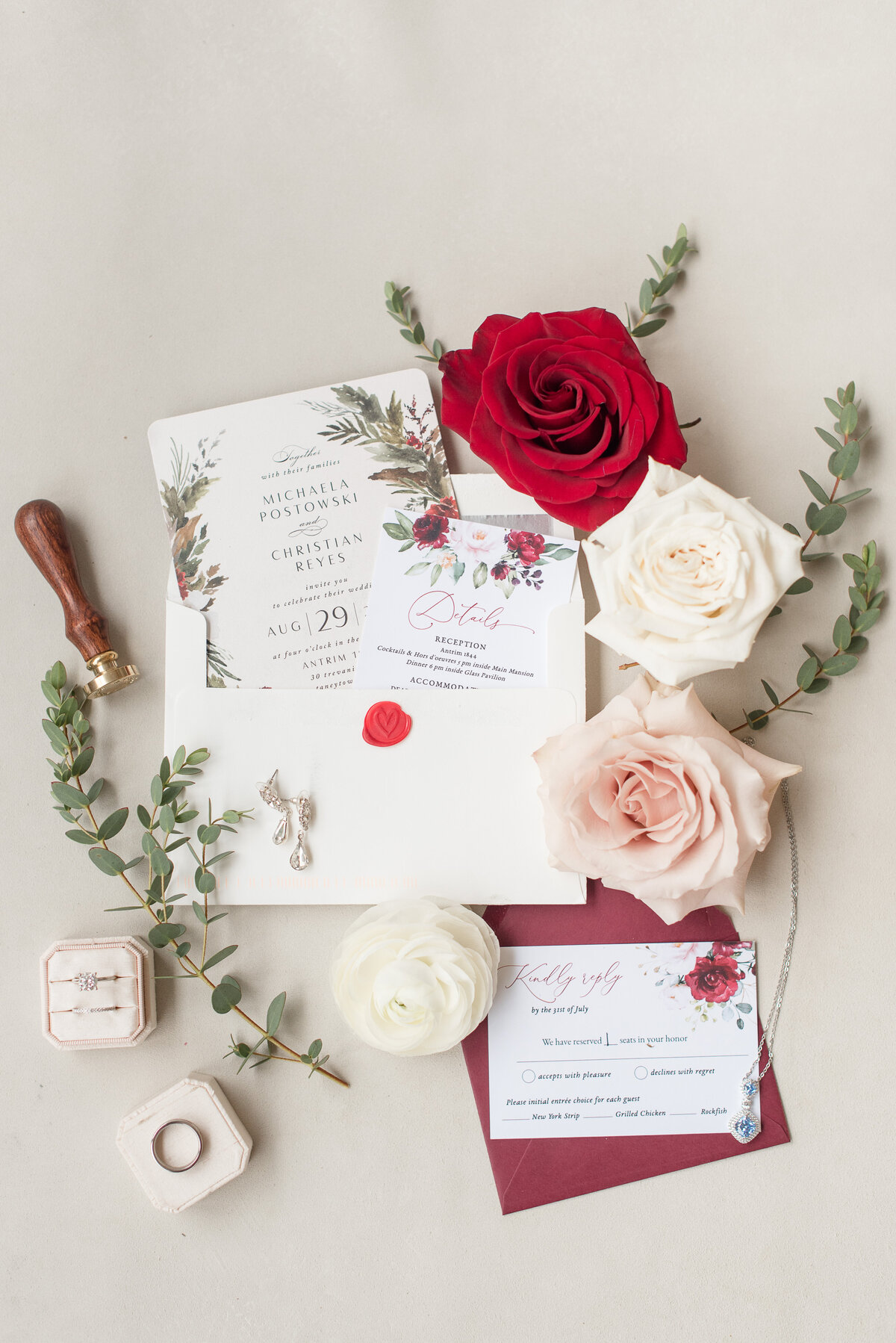 Couples' wedding invitation and wedding rings in boxes displayed on table with roses and eucalyptus.