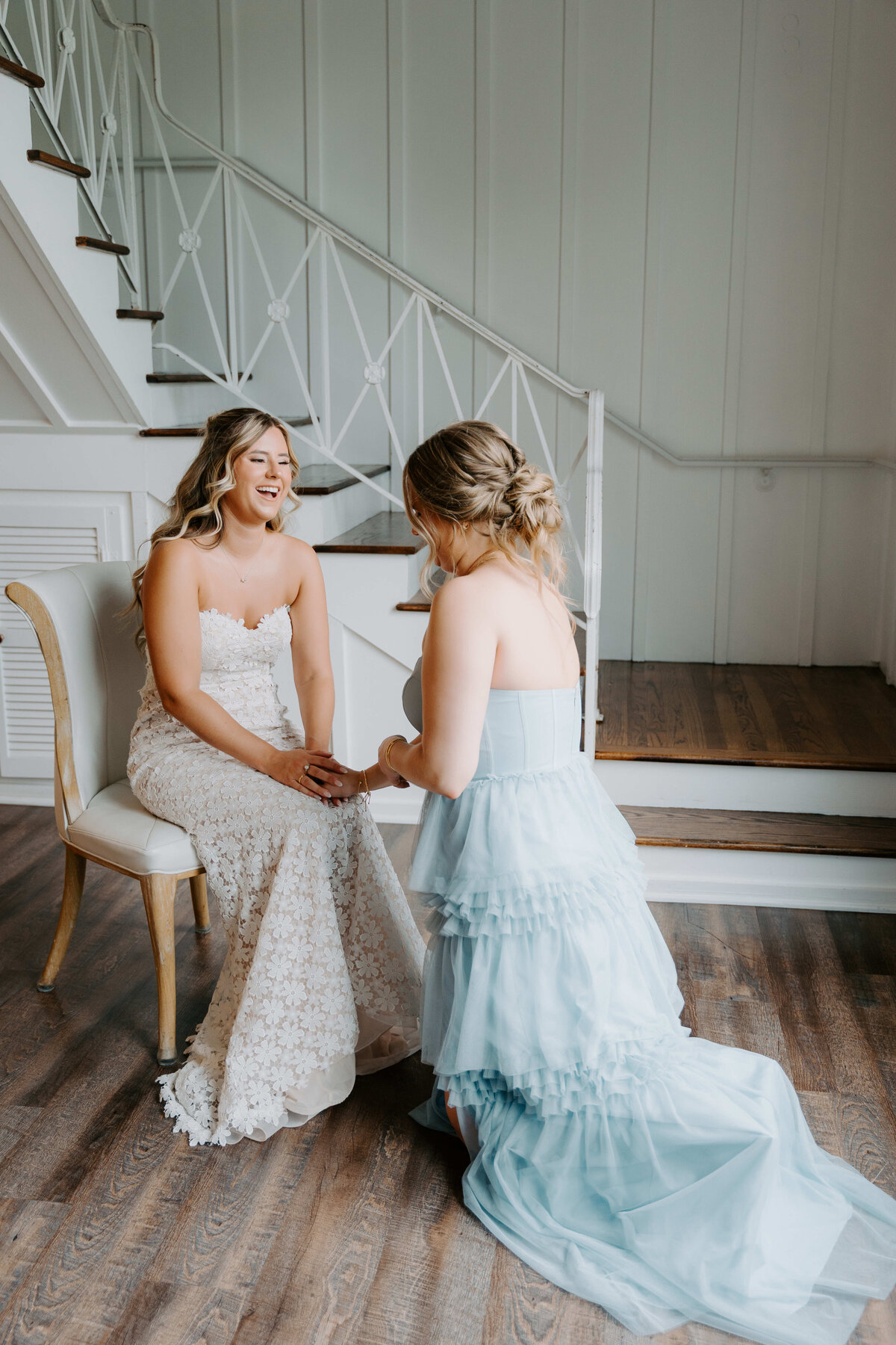 A bride in a lace gown sits on a chair, while an attendant in a light blue dress kneels to adjust her shoe. The setting is a rustic room with stairs, conveying a calm, intimate moment.