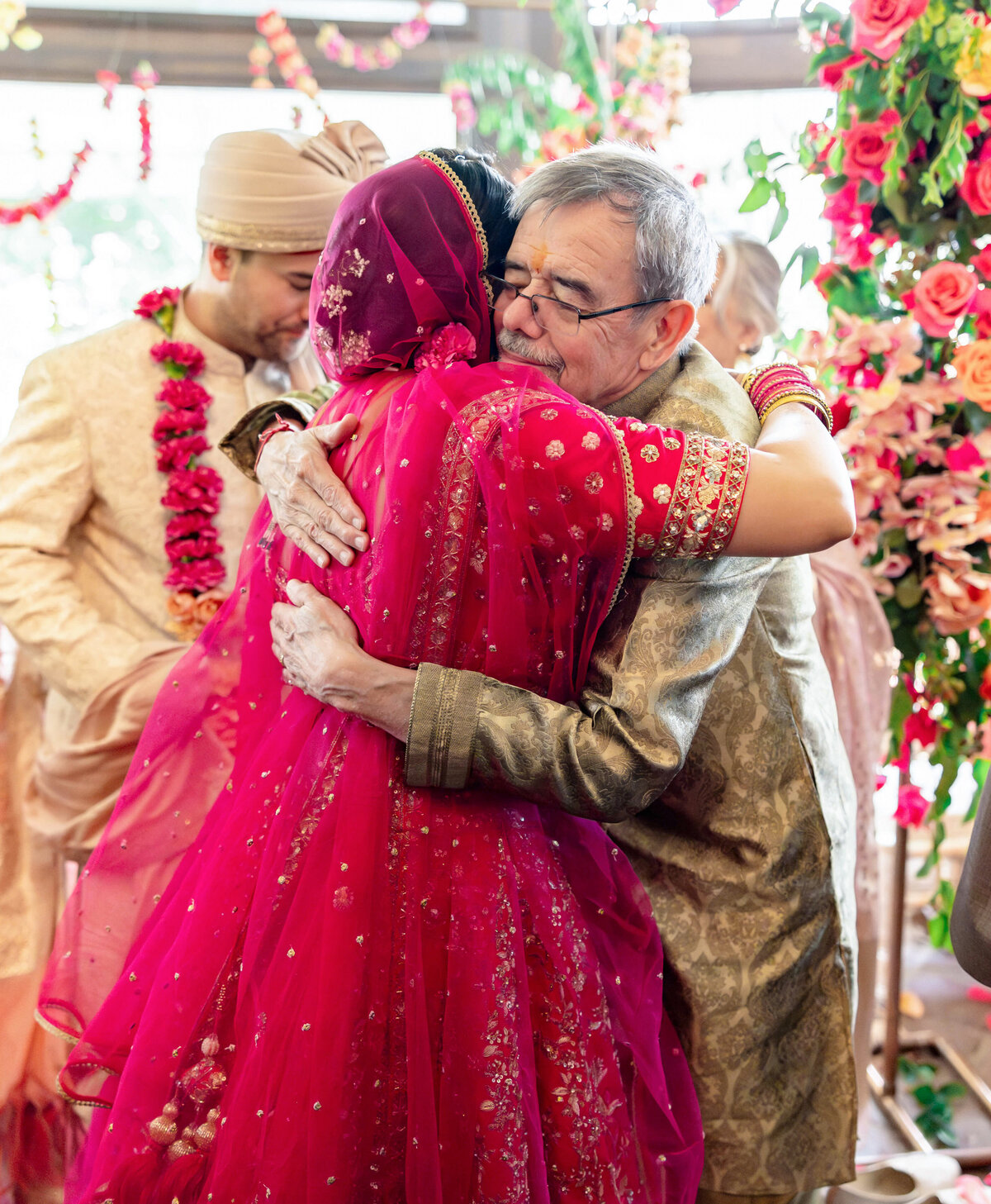 A bride in a vibrant red and gold saree warmly hugs an older man in a gold outfit. In the background, a man in a beige turban and garland watches. The scene is adorned with colorful flowers and festive decorations.