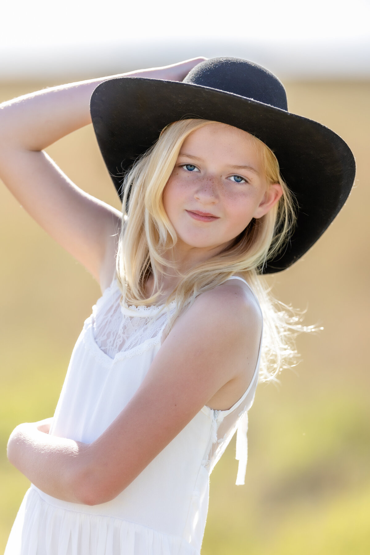 Young girl is wearing a white sleeveless dress with a black cowboy hat and is looking at the camera
