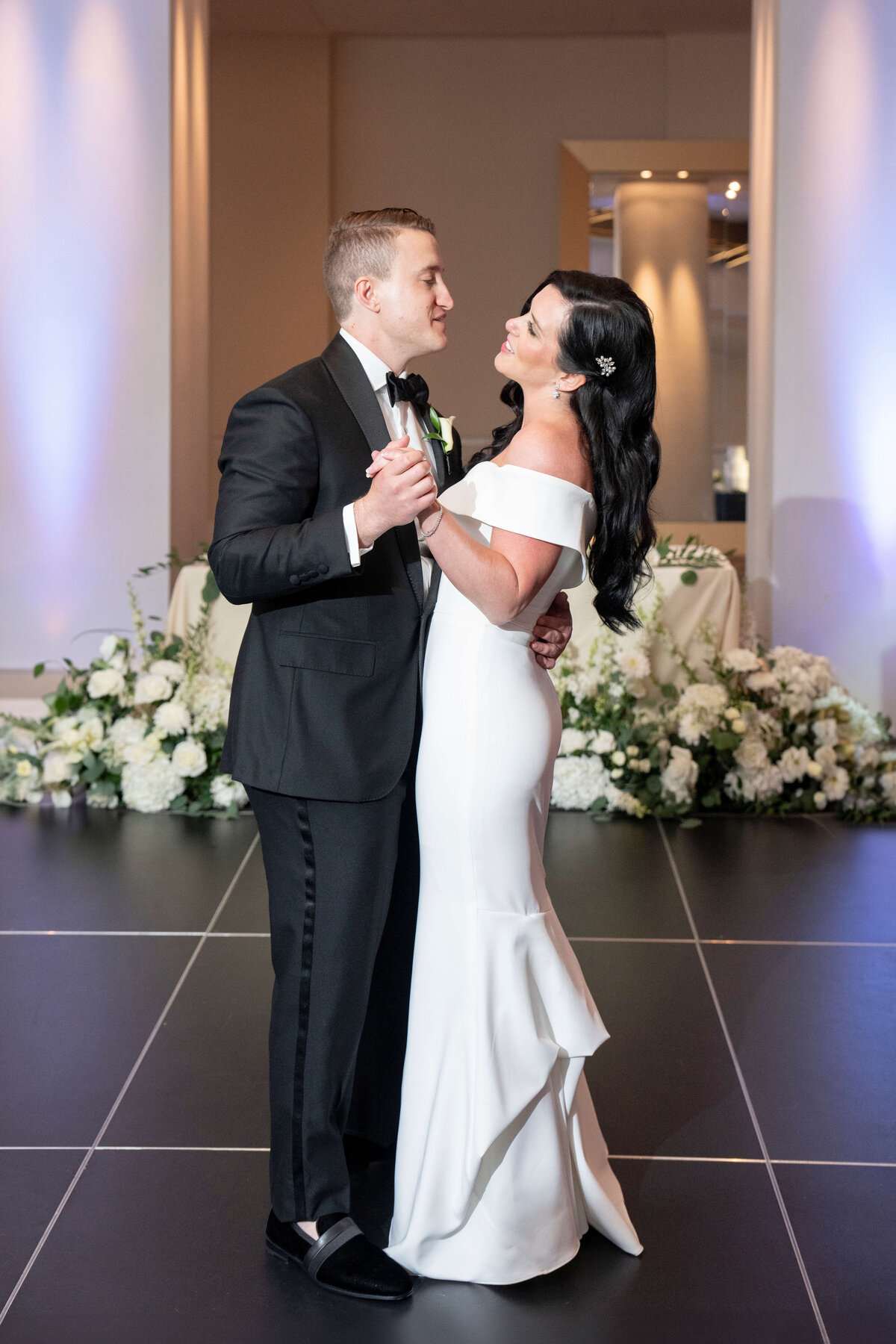 A couple dances together at a wedding reception. The woman is wearing an elegant white gown and the man is in a black tuxedo. They are smiling and surrounded by floral decorations on a black and white checkered dance floor.
