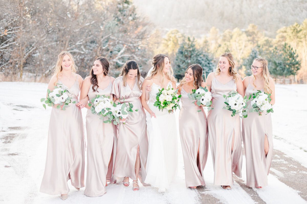 Group of bridesmaids and bride walking arm in arm down a snowy road.