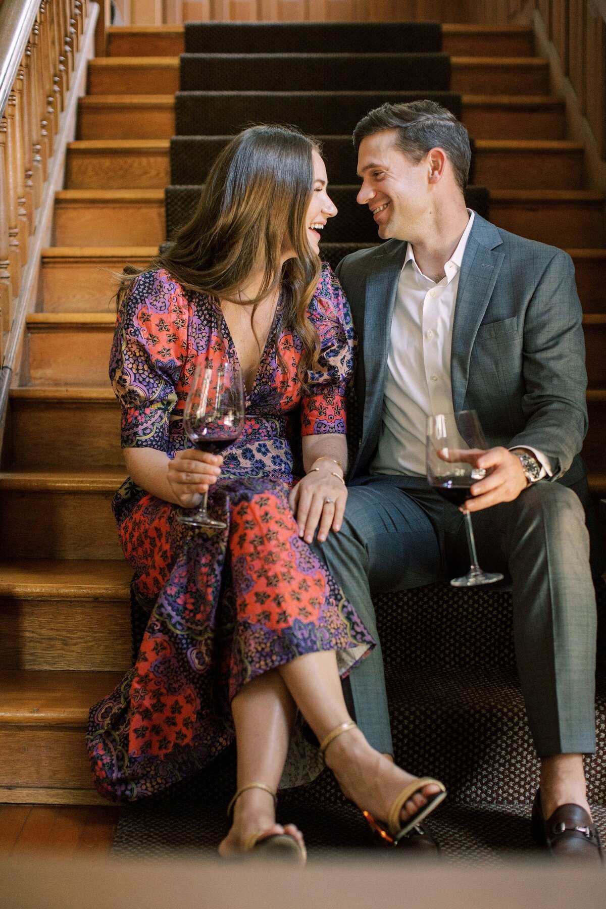 engaged couple smile at each other while sitting on a staircase drinking wine