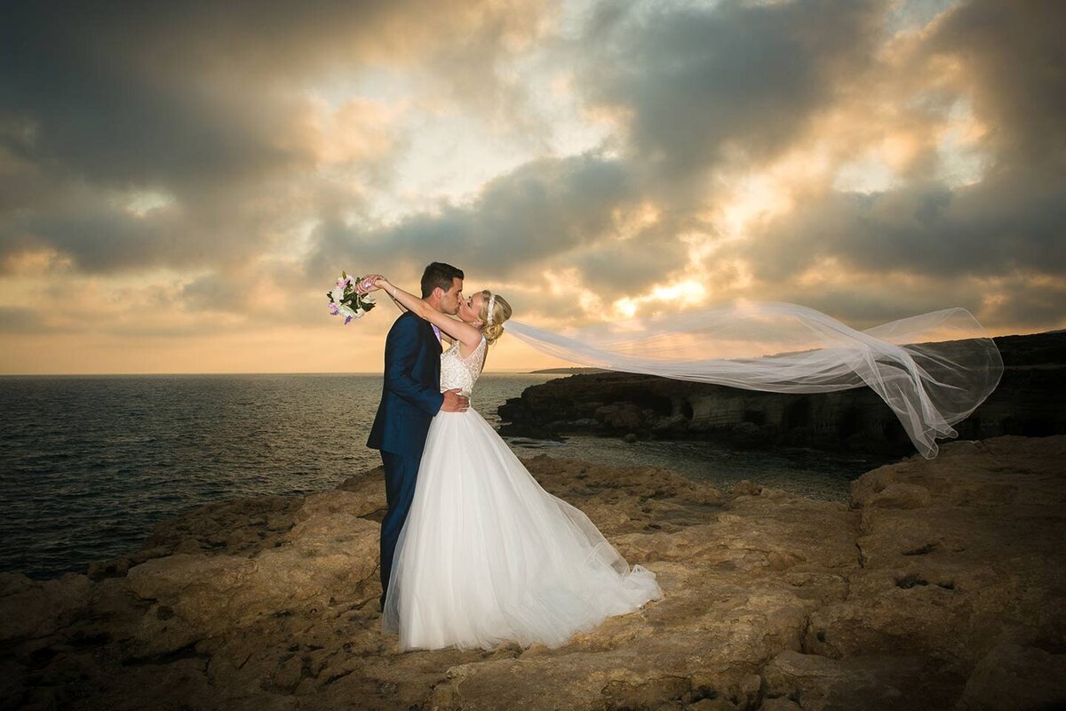 A couple kiss at sunset as the brides veil flies high above in the breeze