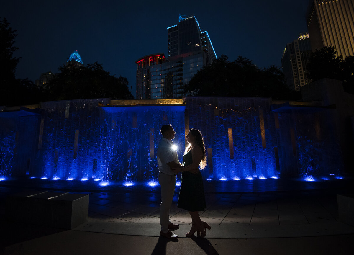 Silhouette-of-a-couple-holding-hands-in-front-of-the-blue-waterfall-in-Romare-Bearden-Park-with-the-city-skyline-in-the-background