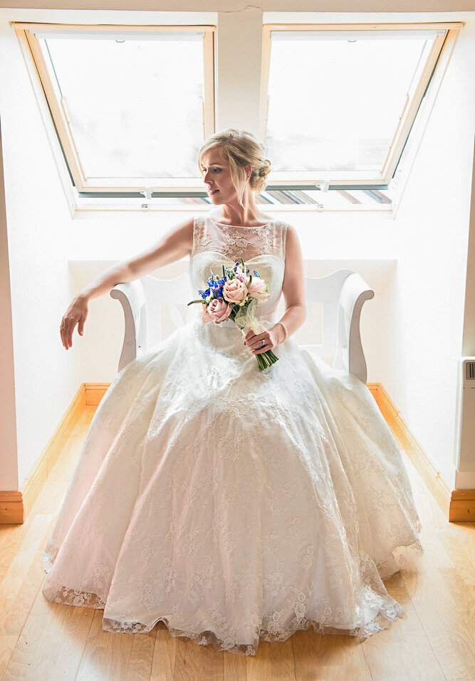 blonde bride wearing an a-line, sweetheart neckline dress sitting on a white seat under window light while holding a pink and purple wedding bouquet