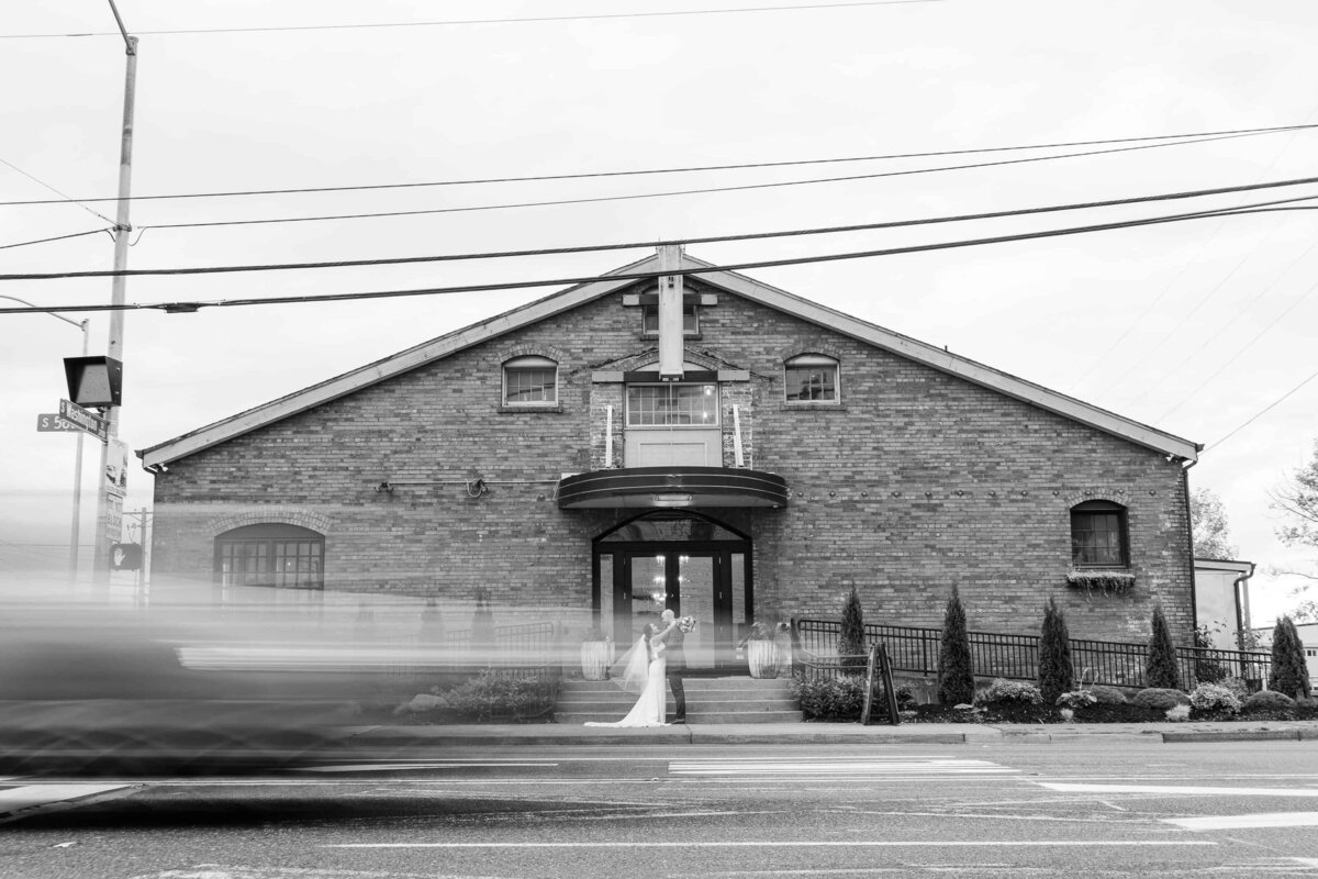 Black and white image of bride and groom, standing in front of The Realm Venue in Tacoma while cars are blurry and wizz by