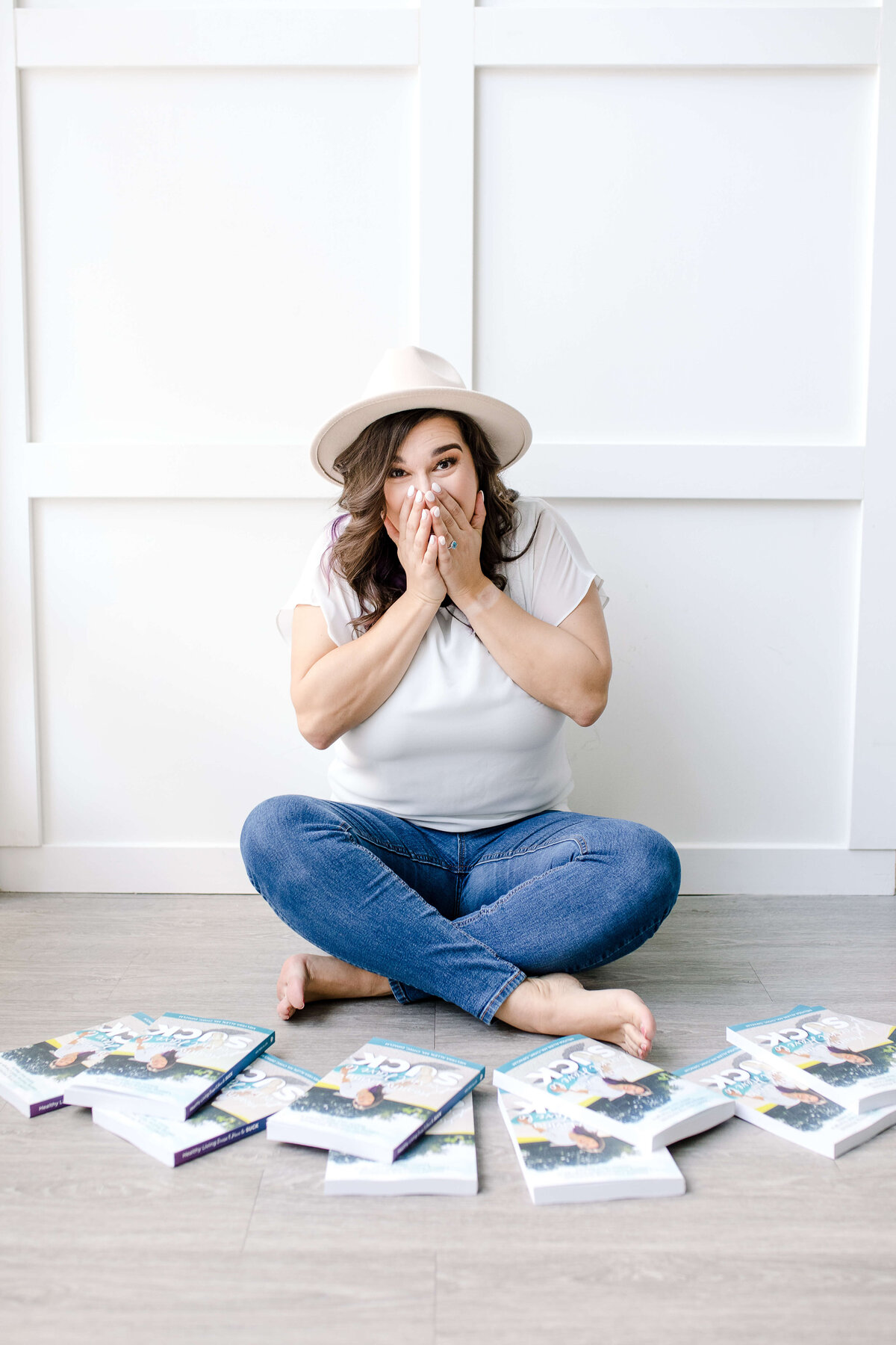 Brand photographer near me photographs woman sitting crisscross on the floor behind a pile of books she has published with her hands to her mouth and surprise for branding photo shoot ideas