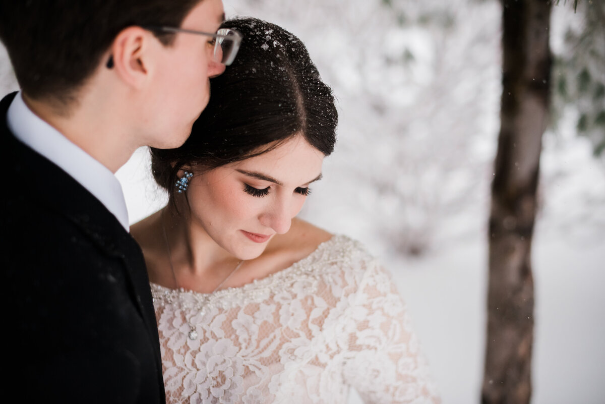 denver wedding photographer captures winter wedding portraits with groom kissing his brides head as she wears a long sleeve lace wedding dress and looks down to her bouquet