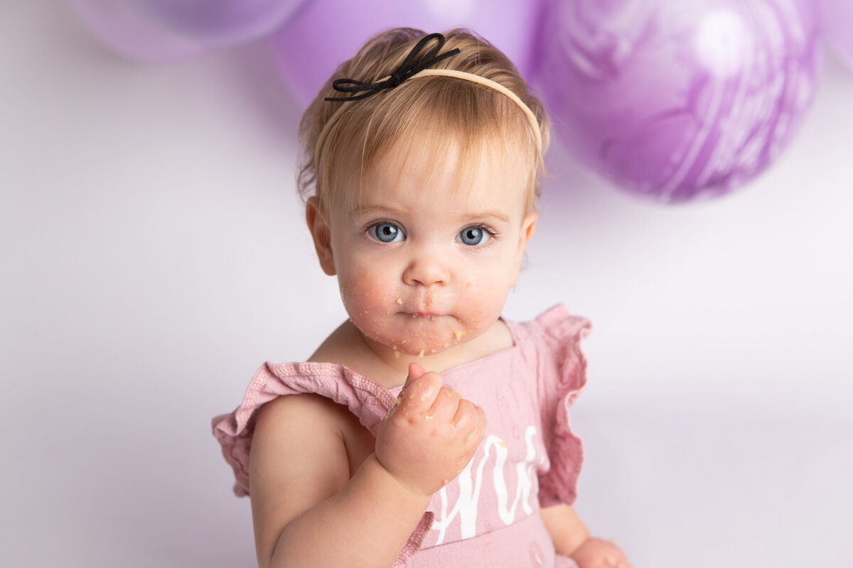Baby girl eating a messy cake with a purple balloon garland