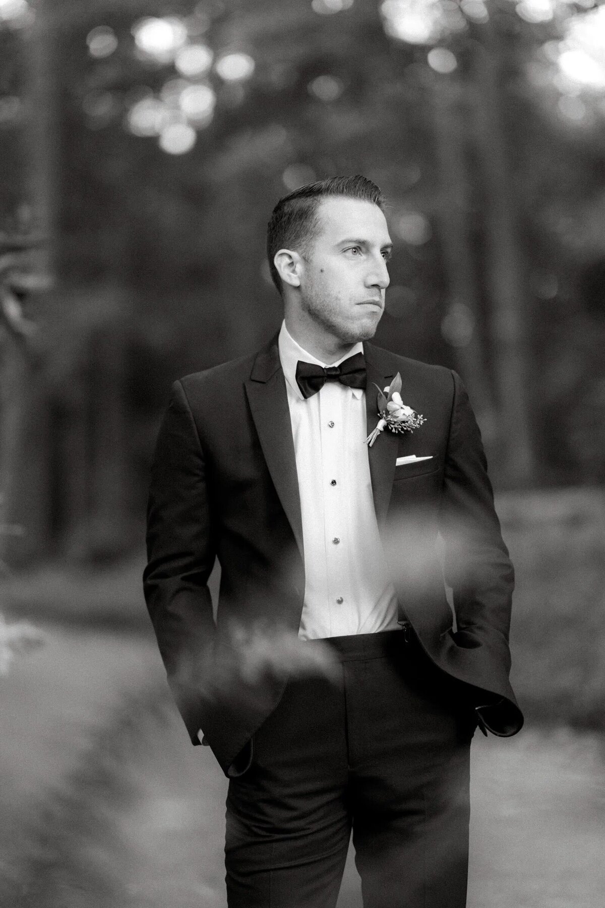 A groom in a black tuxedo and bow tie looking contemplative in a forested path, captured in a black and white portrait.
