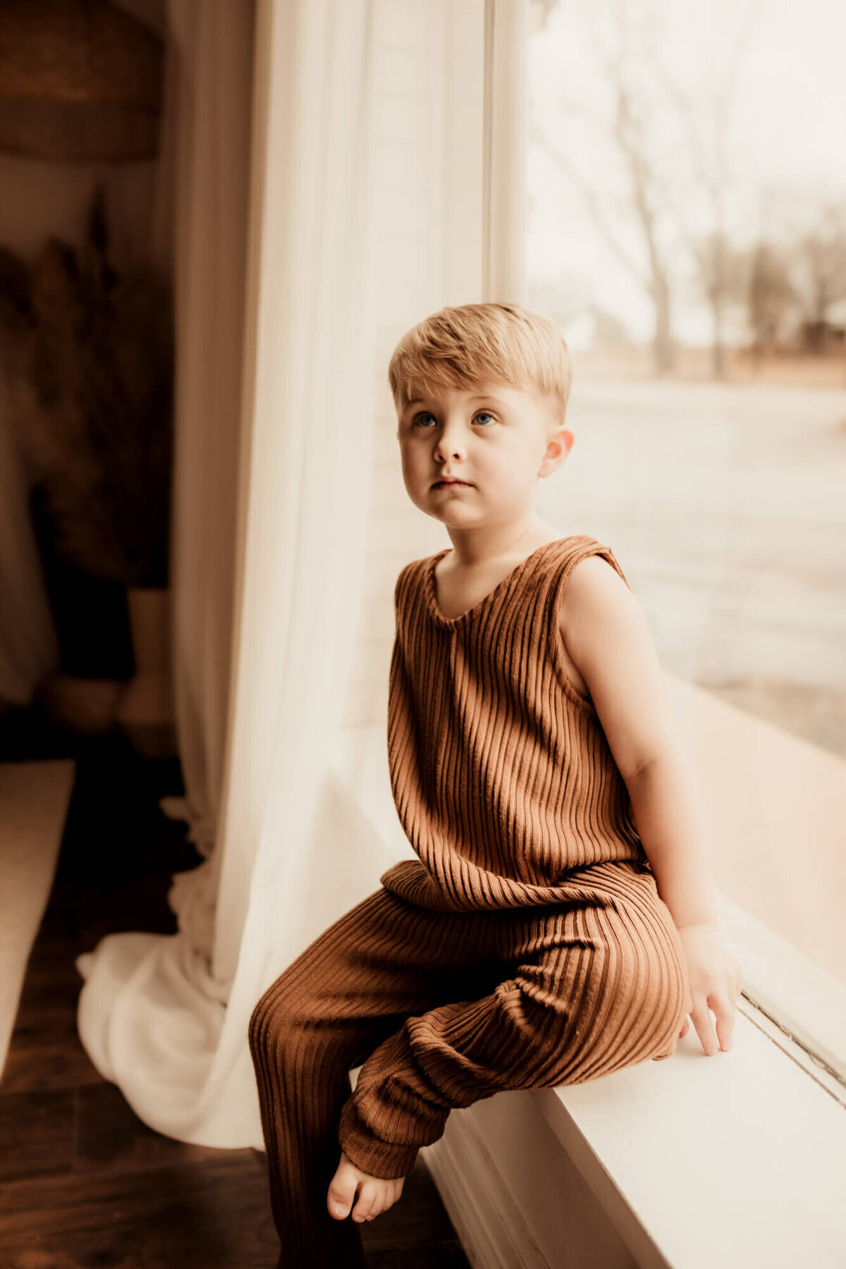 Little boy wearing a brown romper sitting on a window ledge.