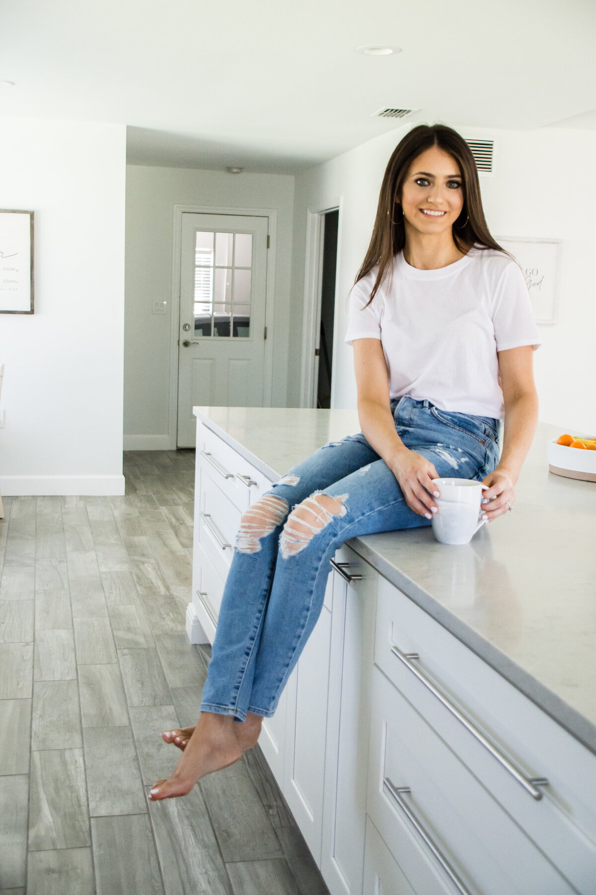 Woman with white coffee mug sits casually on kitchen countertop